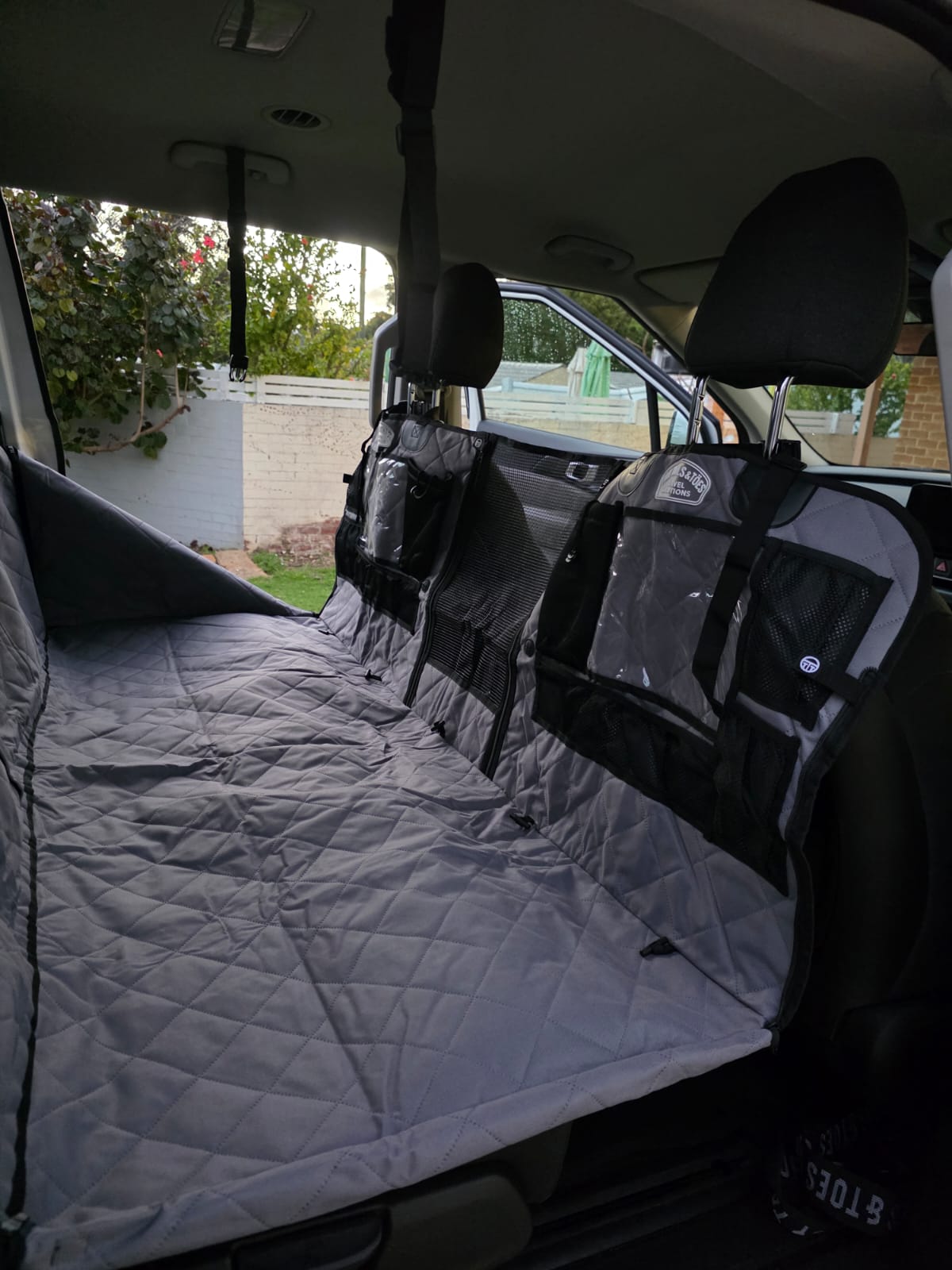 Grey Tidy Traveller quilted back seat car protector and organiser in a Kia Carnival with black harness straps and mesh pockets installed in the back seat of a vehicle.